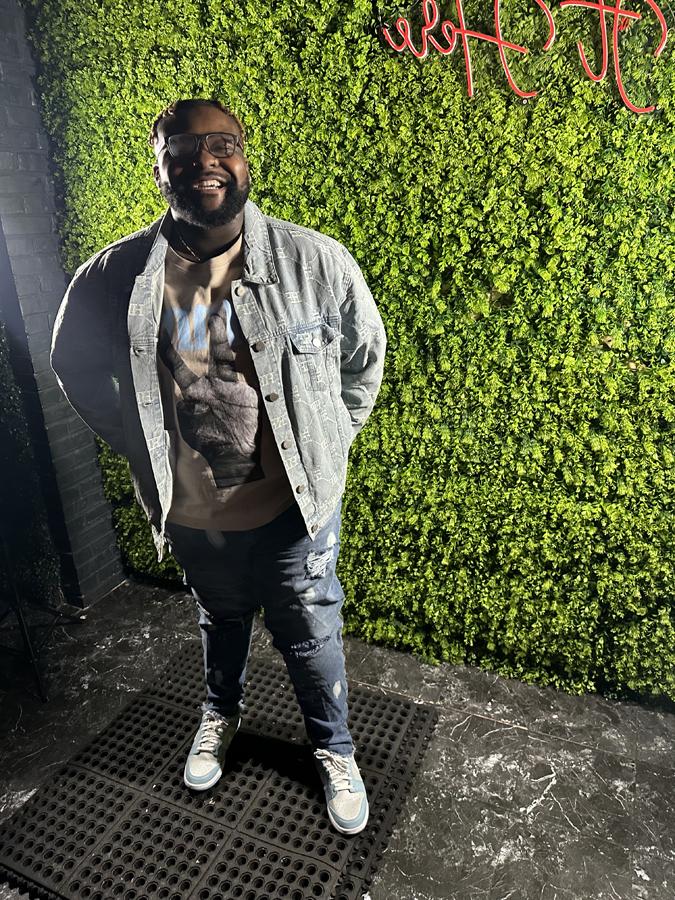 Eric Jones stands in front of a wall with faux greenery and black-painted brink, wearing Nike tennis shoes, jeans and a denim jacket. 