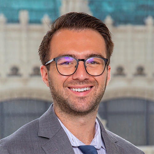 Image of Travis Handler wearing large, 轮, black-framed glasses, a blue and white houndstooth shirt and grey blazer. In the backg轮 is a gothic-style building with a green roof.