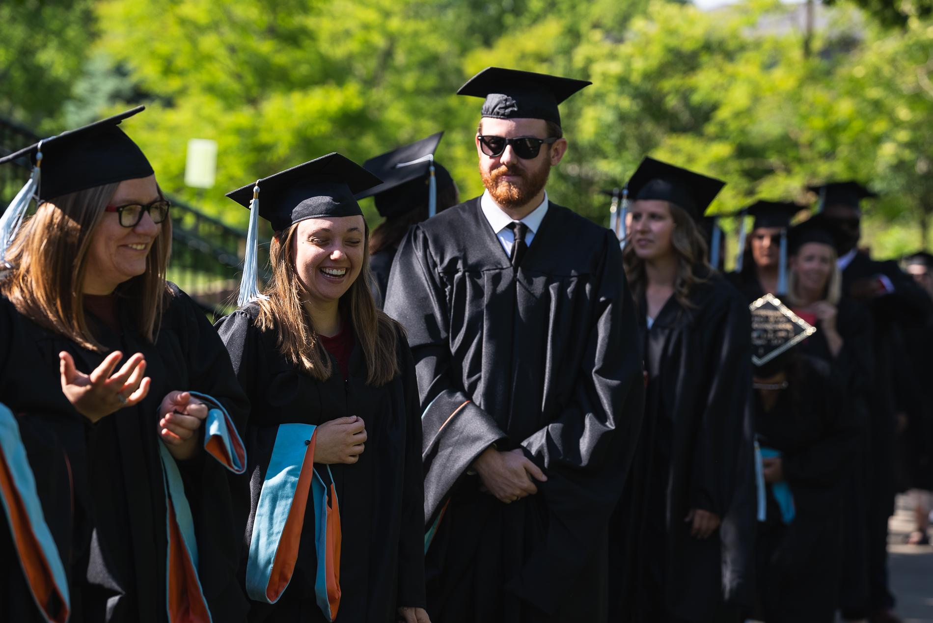 Graduates in black gowns 和 caps, with blue tassels, 在排队参加毕业典礼时开怀大笑.