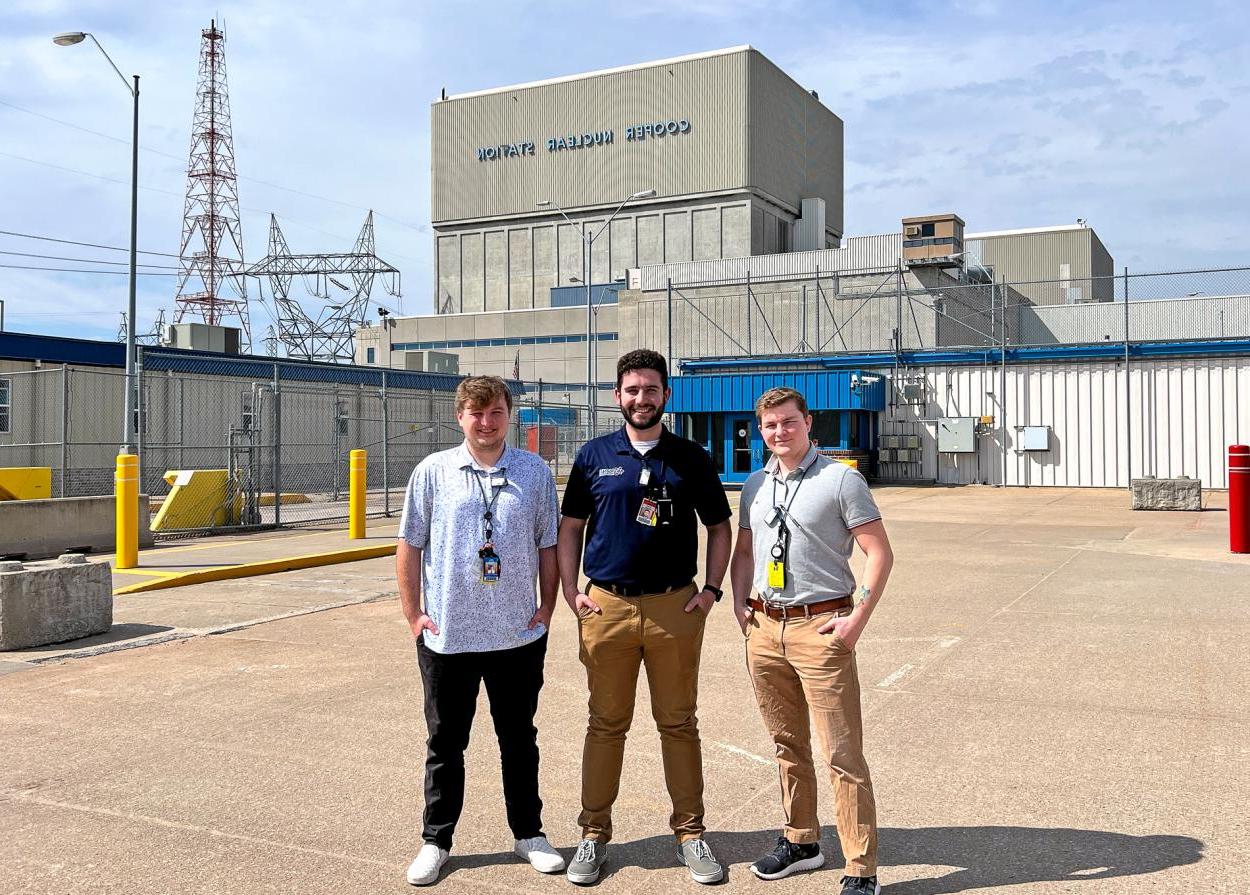 Three men stand in the sunlight of a concrete parking lot, in front of a blocky concrete building bearing the words "Cooper Nuclear Station."