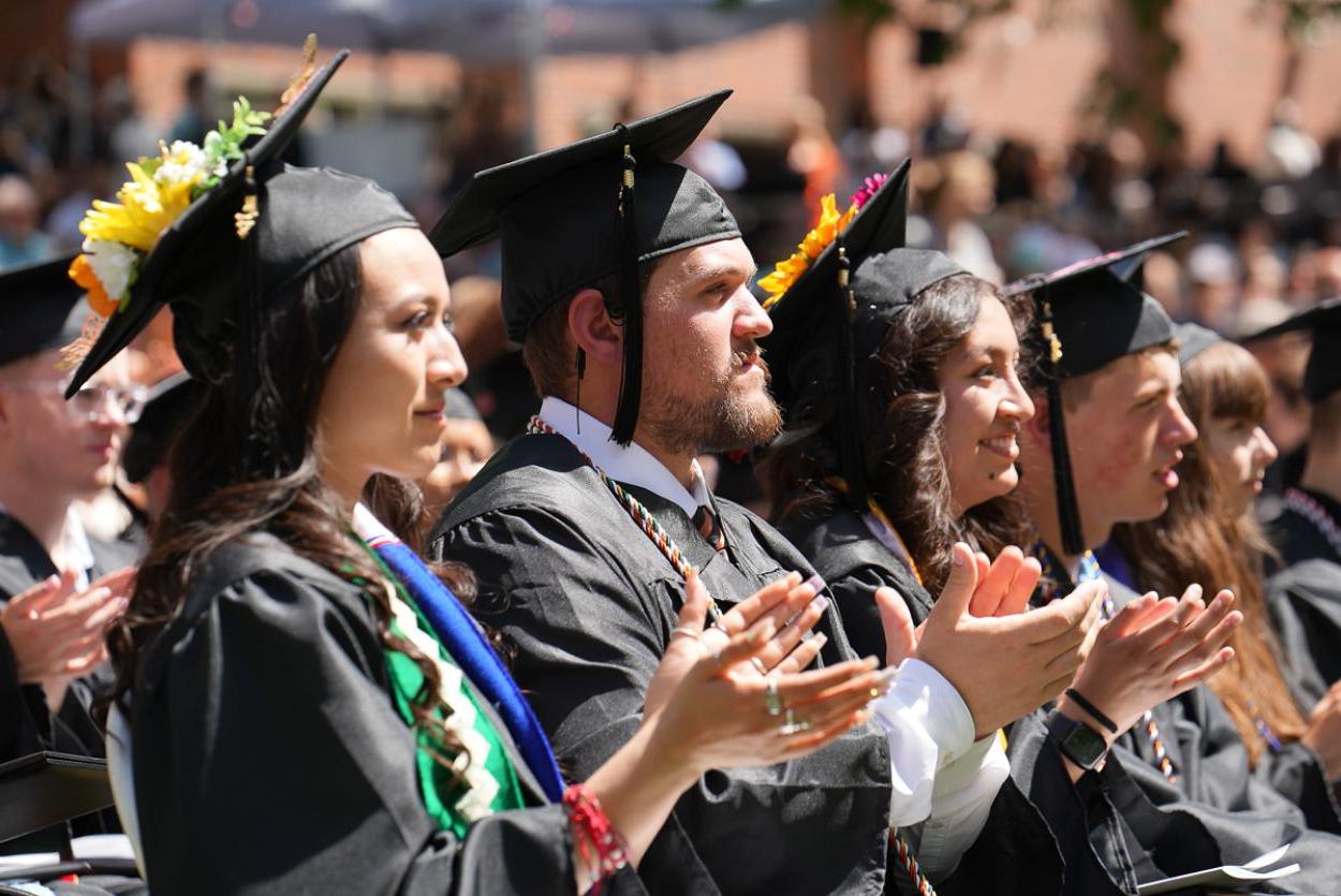 A row of graduates in black caps and gowns clap their hands, with rows of other students and families visible behind them.