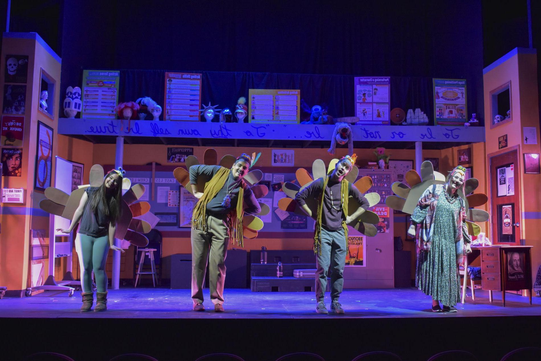 Four actors on a stage lit with purple and yellow lights, 一个穿长裙的女人, two men wearing dark pants, 衬衫和流苏背心, and a second woman in a tshirt, 打底裤和靴子. They're all wearing felt turkey tails and leaning to their right. The stage is set to look like a theater classroom, with props and posters on three walls and the fourth open to the audience.