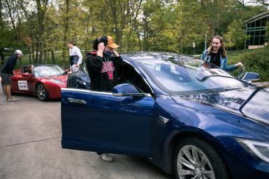 Four Doane students climb into the driver's and passenger's sides of two Tesla vehicles, a blue sedan in the foreground and a sporty red roadster in the background.