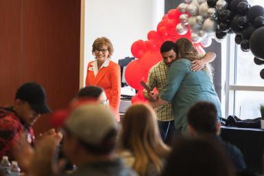 Senior Arian Alai, wearing a green plaid shirt, hugs Jill Kline, wearing a teal jacket, in front of a black, silver and red balloon arch. In the foreground, students and their families are seated, with only Arian and Jill in focus.